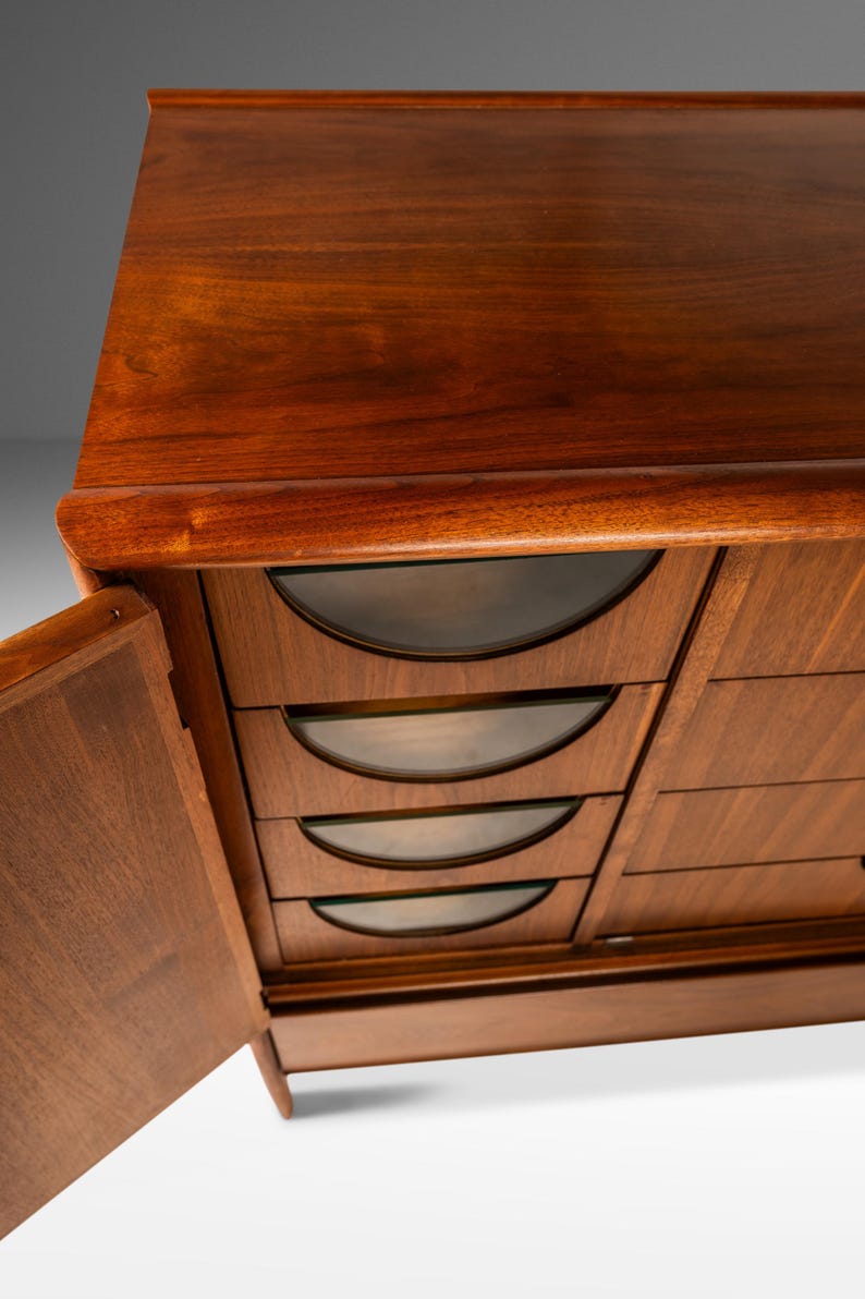 May include: A vintage wooden dresser with six drawers and a cabinet door. The drawers have curved glass fronts and the cabinet door is open.
