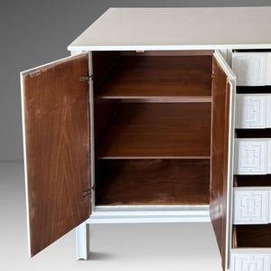 May include: White cabinet with open doors revealing wooden shelves. The cabinet has a white exterior and a series of drawers with a geometric design. The interior of the cabinet is a warm brown color.