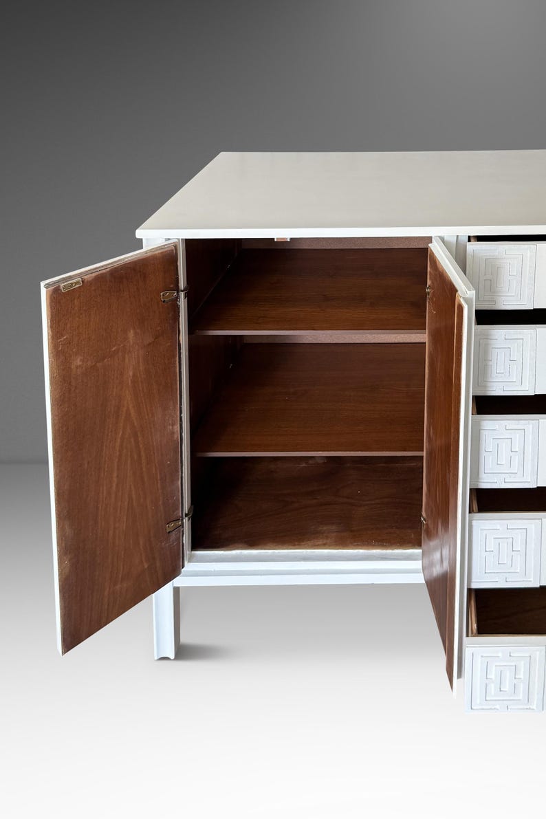 May include: White cabinet with open doors revealing wooden shelves. The cabinet has a white exterior and a series of drawers with a geometric design. The interior of the cabinet is a warm brown color.