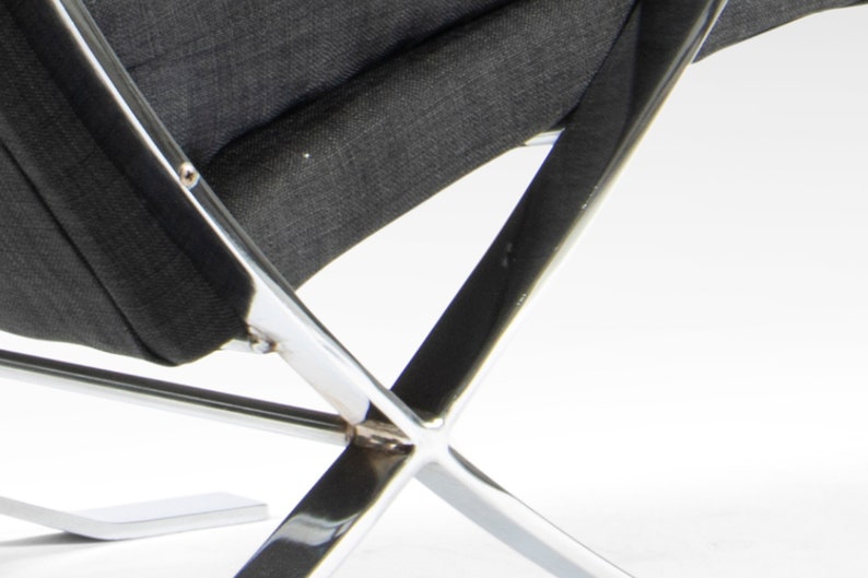 May include: Close-up of the base of a black fabric chair with a chrome frame. The frame is made of four chrome bars that intersect in the center. The chair is on a white surface.