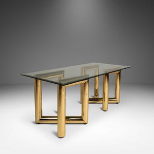 May include: A rectangular glass-top table with a gold-colored metal frame. The table has a minimalist design with thick, cylindrical legs and a clear glass surface. The table is approximately 72 inches long.