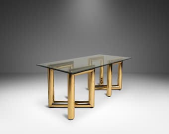 May include: A rectangular glass-top table with a gold-colored metal frame. The table has a minimalist design with thick, cylindrical legs and a clear glass surface. The table is approximately 72 inches long.