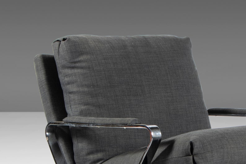 May include: A gray upholstered armchair with a chrome frame. The chair has a high back and a wide seat. The upholstery is a textured fabric with a subtle pattern.