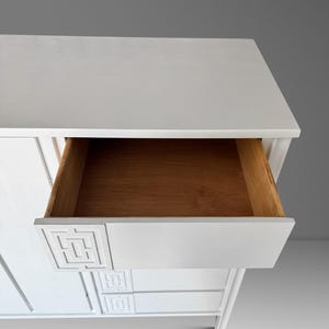 May include: White wooden dresser with an open drawer revealing a natural wood interior. The drawer front features a geometric design. The dresser has a clean, modern aesthetic, with a minimalist design and a neutral color palette.