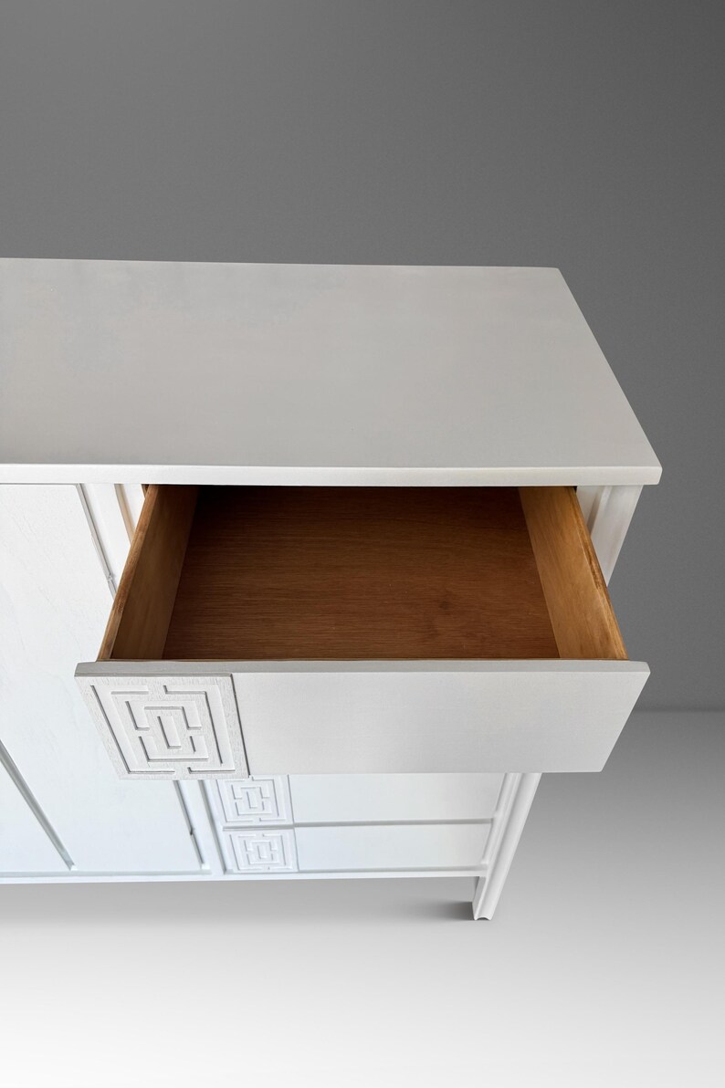 May include: White wooden dresser with an open drawer revealing a natural wood interior. The drawer front features a geometric design. The dresser has a clean, modern aesthetic, with a minimalist design and a neutral color palette.