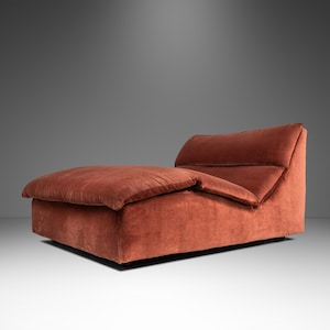 May include: A modern, low-profile chaise lounge in a warm, reddish-brown hue. The plush, upholstered seat and back offer a comfortable design. The chaise is approximately 72 inches long and 36 inches wide, with a low back for support.