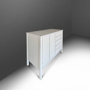 May include: A white wooden cabinet with a rectangular top, two doors, and four drawers. The cabinet has a simple, modern design with clean lines and a light finish. The cabinet is set against a gray background.