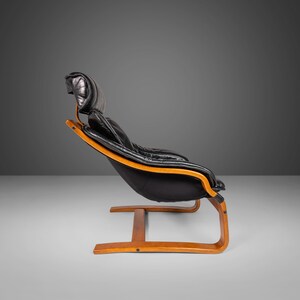 May include: A black leather armchair with a wooden frame. The chair has a curved back and a low seat. The chair is on a white background.