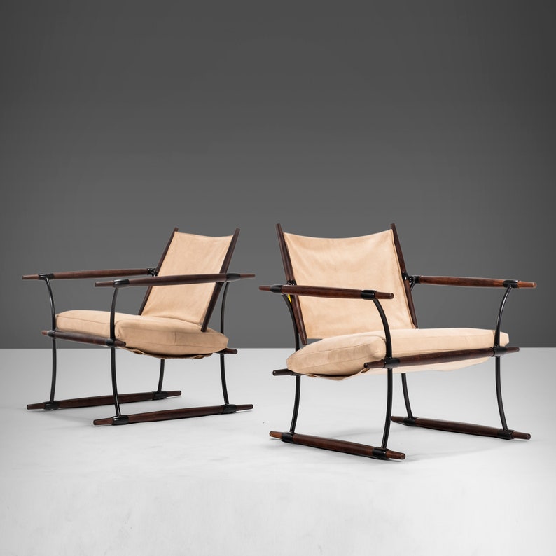 以下が含まれることがあります： 黒い金属フレームとダークウッドのアクセントが付いた、ミッドセンチュリーモダンなラウンジチェア2脚。椅子はベージュのファブリック張りで、白い表面に置かれています。