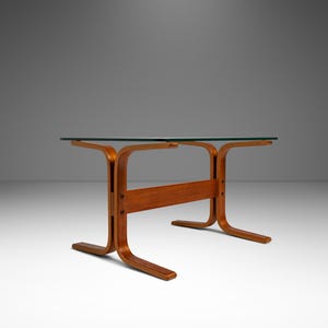 May include: A mid-century modern coffee table with a clear glass top and a curved wooden base. The base is made of light brown wood and has a unique, sculptural design. The table is approximately 48 inches long and 18 inches high.
