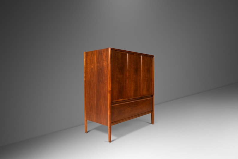 May include: A mid-century modern cabinet with a wood grain finish. The cabinet has two doors and a single drawer. The cabinet is made of solid wood and has a warm brown color.