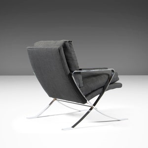 May include: A gray fabric armchair with a chrome frame. The chair has a curved back and a wide seat. The chrome frame is made up of thin, angled pieces that create a unique and modern design.