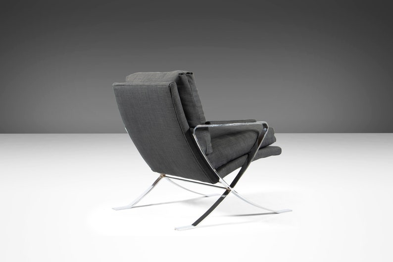 May include: A gray fabric armchair with a chrome frame. The chair has a curved back and a wide seat. The chrome frame is made up of thin, angled pieces that create a unique and modern design.