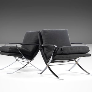 May include: Two black fabric chairs with chrome frames. The chairs have a simple design with a low back and a wide seat. The chairs are positioned on a white surface.