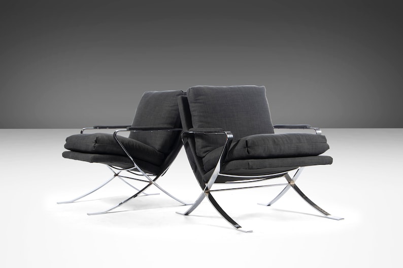 May include: Two black fabric chairs with chrome frames. The chairs have a simple design with a low back and a wide seat. The chairs are positioned on a white surface.
