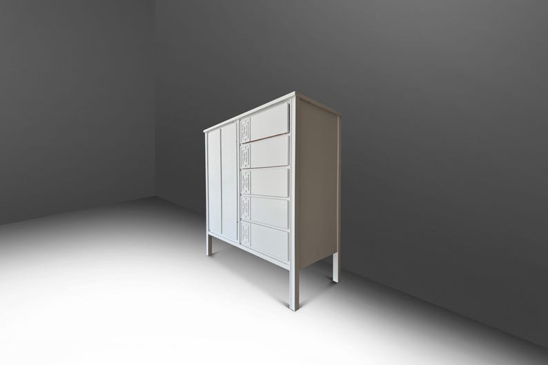 May include: A white cabinet with a vertical panel on the left and five drawers on the right. The drawers have decorative rectangular panels. The cabinet stands on four thin legs. The background is a gradient of gray.