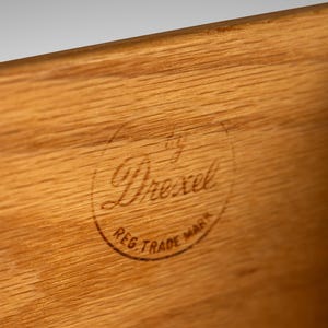 May include: Close-up of a wood surface with a circular, brown, stamped logo that reads "by Drexel Reg. Trade Mark".