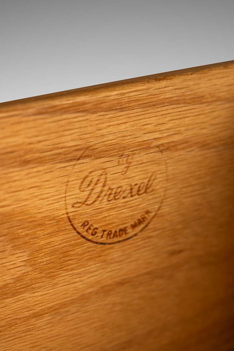 May include: Close-up of a wood surface with a circular, brown, stamped logo that reads "by Drexel Reg. Trade Mark".