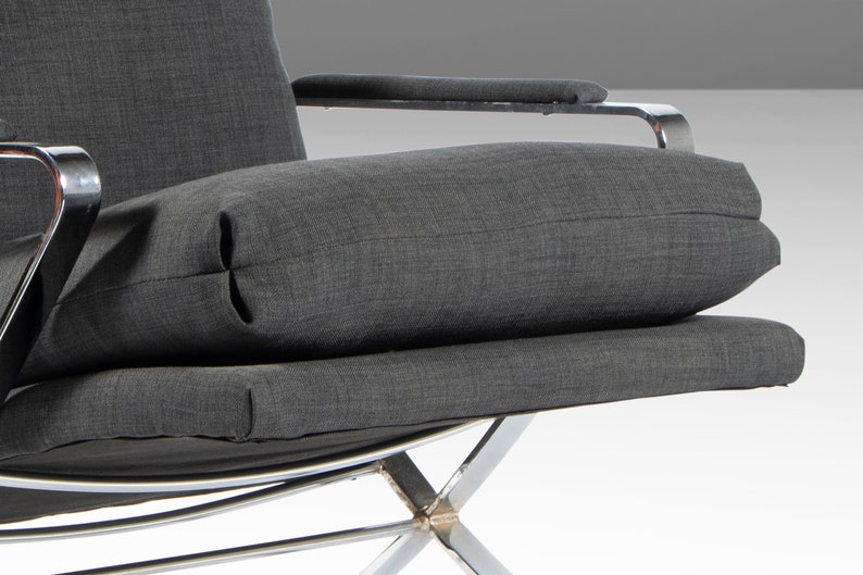 May include: A gray fabric armchair with a chrome frame. The chair has a thick, gray fabric cushion and a smaller, gray fabric pillow. The chair is angled slightly to the right.