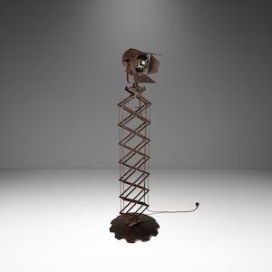 May include: A vintage-style floor lamp with a rusty brown finish. The lamp features an adjustable scissor arm and a spotlight head. The base is a dark, circular shape. The lamp is designed for illumination.