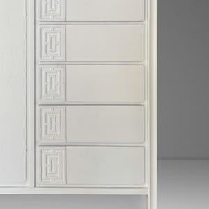 May include: A white wooden dresser with five drawers and a cabinet door. Each drawer features a recessed geometric design. The dresser has a clean, modern aesthetic, with a simple frame and legs.