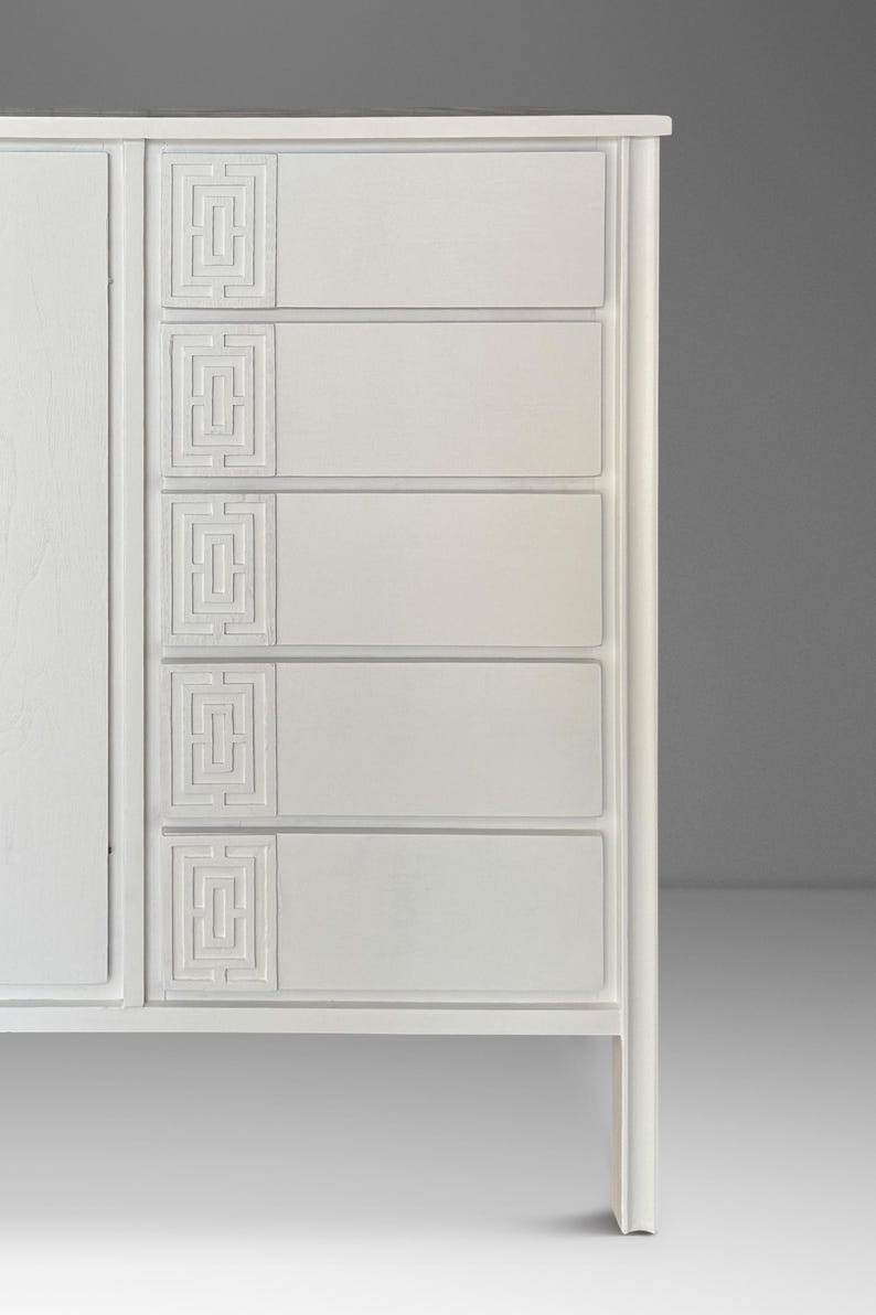 May include: A white wooden dresser with five drawers and a cabinet door. Each drawer features a recessed geometric design. The dresser has a clean, modern aesthetic, with a simple frame and legs.