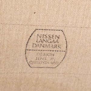 以下が含まれることがあります： 「NISSEN LANGAA DANMARK DESIGN Jens H. QUISTGAARD」と黒文字で書かれた茶色の布製のラベルのクローズアップ。