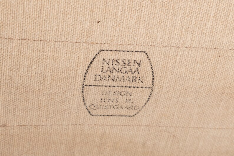 以下が含まれることがあります： 「NISSEN LANGAA DANMARK DESIGN Jens H. QUISTGAARD」と黒文字で書かれた茶色の布製のラベルのクローズアップ。