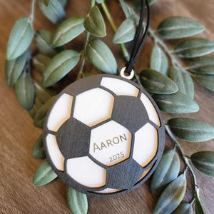 May include: A soccer ball-shaped ornament with a black and white design. The ornament has the name "Aaron" and the year "2025" engraved on it. A black ribbon is attached for hanging. The ornament is surrounded by green leaves.