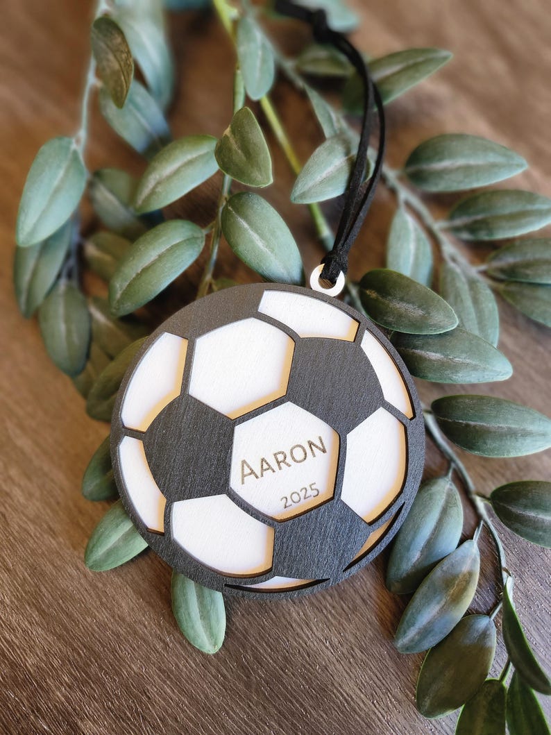 May include: A soccer ball-shaped ornament with a black and white design. The ornament has the name "Aaron" and the year "2025" engraved on it. A black ribbon is attached for hanging. The ornament is surrounded by green leaves.