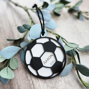 May include: A black and white soccer ball ornament with the name "Aaron" and the year "2025" printed on it. The ornament is hanging from a black string and is surrounded by green and blue leaves.