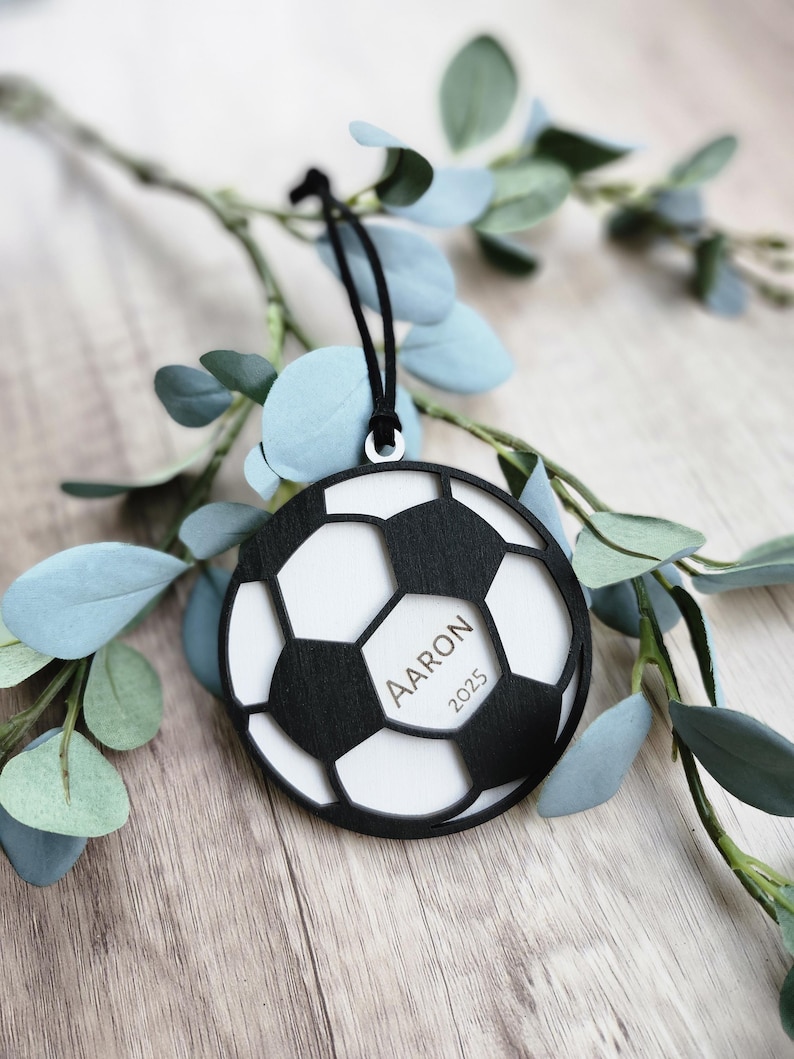 May include: A black and white soccer ball ornament with the name "Aaron" and the year "2025" printed on it. The ornament is hanging from a black string and is surrounded by green and blue leaves.