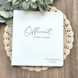 May include: A white rectangular binder with black text that reads Officiant, HENRY ALBERT, LUKE & MAKENNA, and 04.24.2027. The cover features a smooth, matte finish with stitched edges, providing a clean and professional appearance for documents.