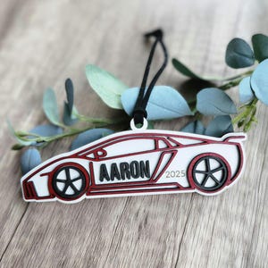 May include: A white and red car-shaped ornament with the name "AARON" and the year "2025" in black. The ornament has a black string for hanging. The car has black wheels and red accents. The background includes greenery and a wooden surface.