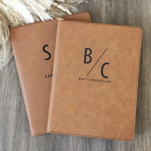 May include: Two tan leather journals with black lettering. One journal has the letter "S" and the word "SAP" printed on it. The other journal has the letters "B/C" and the name "BRETT COUNSELLER" printed on it. The journals are on a wooden surface.