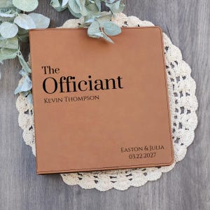 May include: A tan rectangular binder with black text that reads The Officiant, KEVIN THOMPSON, and EASTON & JULIA 03.22.2027. The cover has a smooth, leather-like texture with stitched edges, presented as a functional document holder.