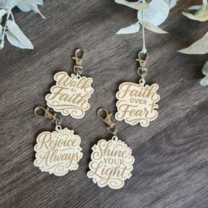 May include: Four wooden keyrings with motivational quotes. Each keyring features a unique design with phrases such as "Walk by Faith," "Faith over Fear," "Rejoice Always," and "Shine your Light." The keyrings are attached to silver-coloured clasps.