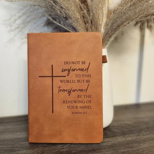 Puede incluir: Un diario de cuero marrón con el texto "DO NOT BE conformed TO THIS WORLD, BUT BE transformed BY THE RENEWING OF YOUR MIND. ROMANS 12:2" y una cruz. El diario se muestra frente a un jarrón blanco con plantas secas.