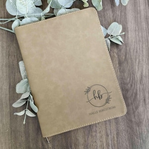Puede incluir: Diario de cuero sintético color beige con un diseño de corona floral en relieve alrededor de las iniciales "HB" y el nombre "HALLE BERGSTROM". El diario está cerrado y tiene un diseño simple y elegante. Hay vegetación decorativa en el fondo.