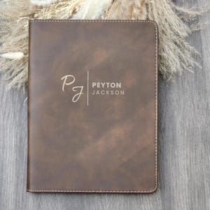 May include: A brown leather-like journal or notebook with the name "Peyton Jackson" printed on the cover. The initials "PJ" are also displayed in a cursive font. The notebook is rectangular with a stitched border.