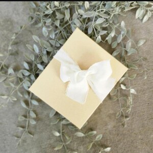 May include: A square, light beige gift box tied with a white, frayed-edge ribbon bow. The box is centered in the image, with soft green leafy sprigs in the background, creating a delicate, natural aesthetic. The box is likely for a small gift.