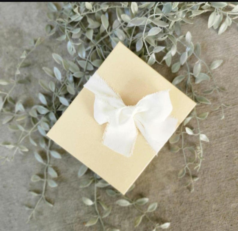 May include: A square, light beige gift box tied with a white, frayed-edge ribbon bow. The box is centered in the image, with soft green leafy sprigs in the background, creating a delicate, natural aesthetic. The box is likely for a small gift.