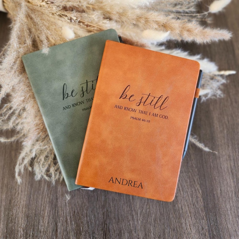 May include: Two leather-bound journals, one sage green and one orange, with the words "be still" and "AND KNOW THAT I AM GOD. PSALM 46:10" printed on the covers. The orange journal is personalized with the name "ANDREA".