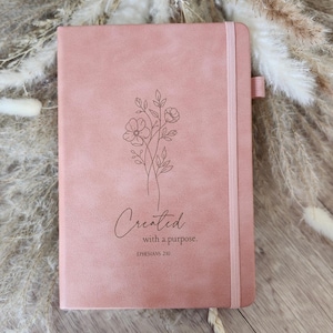 May include: A dusty rose notebook with a vertical elastic closure and a pen loop on the side. The front cover features a black line art illustration of wildflowers and the text "Created with a purpose. EPHESIANS 2:10" printed in black ink.