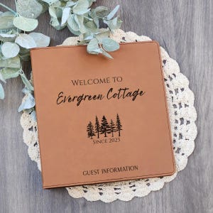 May include: A brown leather-bound guest information book with the text "WELCOME TO Evergreen Cottage" and "GUEST INFORMATION". The cover also features a tree graphic and the text "SINCE 2023". The book rests on a white lace doily.