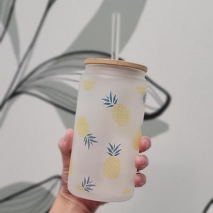 May include: A frosted glass water bottle with a bamboo lid and a pineapple print. The bottle is white with yellow pineapples and blue leaves.