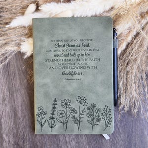 May include: A sage green journal featuring a black floral line art design at the bottom and the text: So then, just as you received Christ Jesus as Lord, continue to live your lives in him, rooted and built up in him, Colossians 2:6-7.