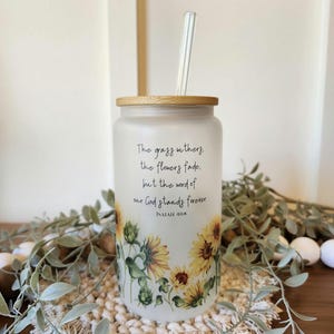 May include: White frosted glass tumbler with a bamboo lid and a straw. The tumbler features a floral design with sunflowers and green leaves. The text "The grass withers, the flowers fade, but the word of our God stands forever, Isaiah 40:8" is printed on the tumbler.