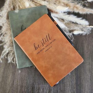 May include: Two notebooks, one olive green and one tan, are displayed. The tan notebook has the words "be still" and "AND KNOW THAT I AM GOD. PSALM 46:10" printed on the cover. A black pen rests on the tan notebook.