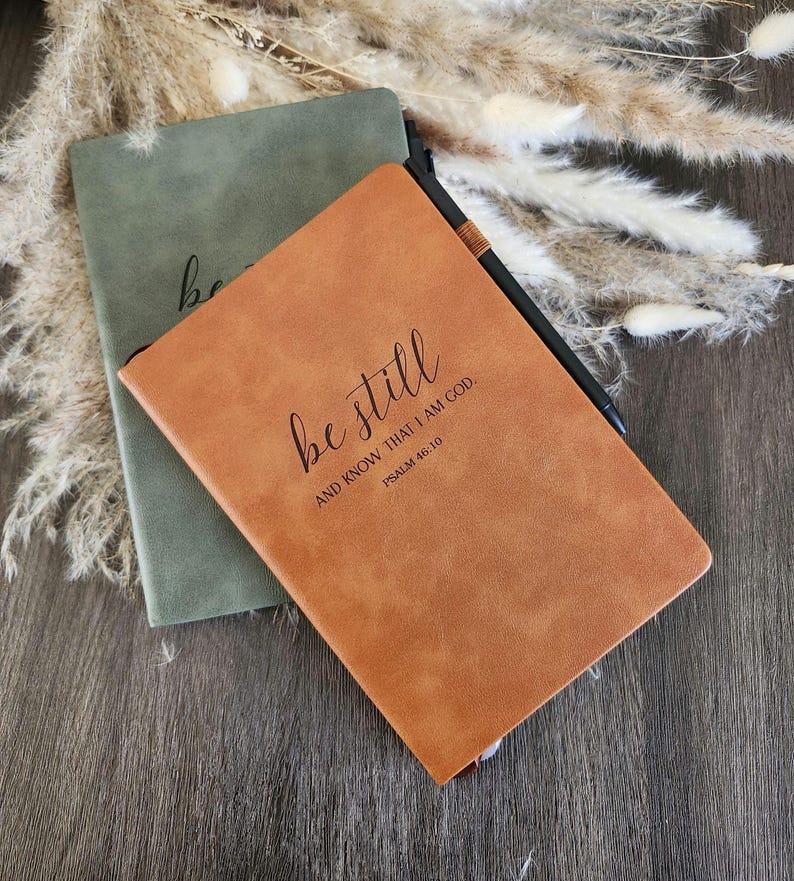 May include: Two notebooks, one olive green and one tan, are displayed. The tan notebook has the words "be still" and "AND KNOW THAT I AM GOD. PSALM 46:10" printed on the cover. A black pen rests on the tan notebook.